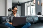 Young woman video calling using a laptop sitting on a sofa wearing earphones