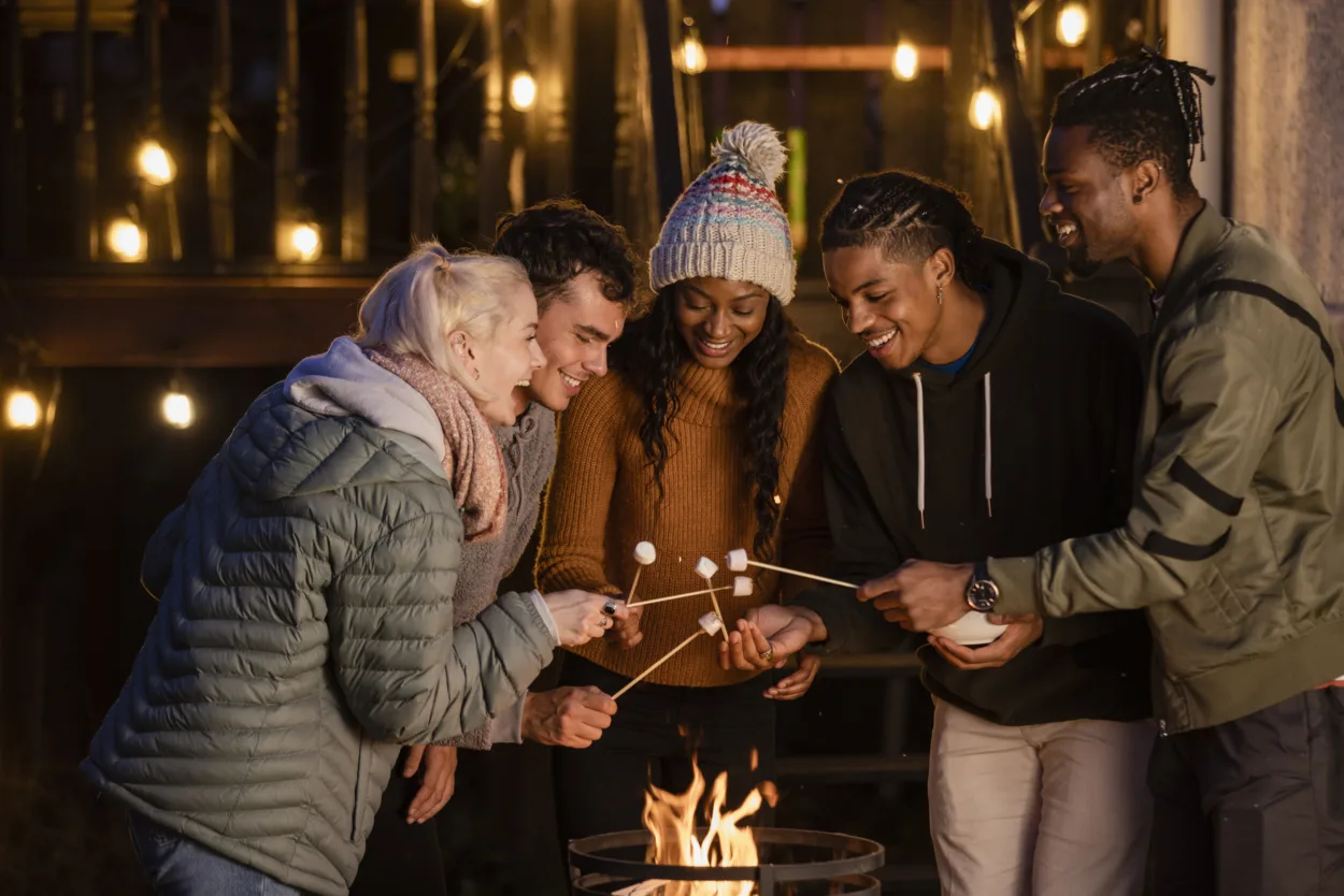 A group of young adults friends are standing around a fire in the dark, smiling. They are heating up the marshmallows they have on sticks.