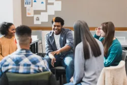 During a group therapy session, a teenage boy gestures as he talks about something with the people in the group. An attentive female therapist facilitates the meeting.