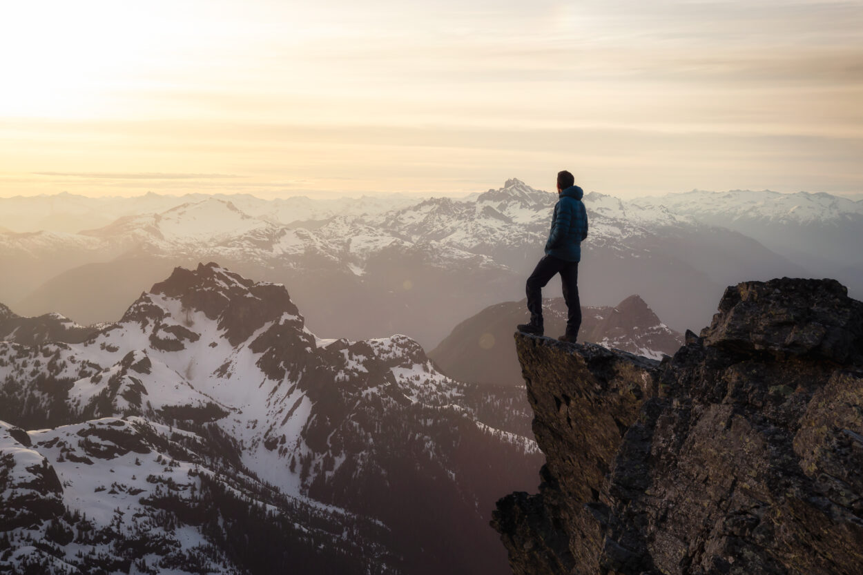 Fantasy Adventure Composite with a Man on top of a Mountain Cliff with Dramatic Landscape in Background during Sunset or Sunrise. Landscape from British Columbia, Canada.