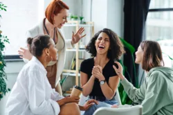 happy psychologist gesturing near excited multiracial woman laughing with closed eyes during motivation session with multiethnic girlfriends, understanding, support and mental health concept