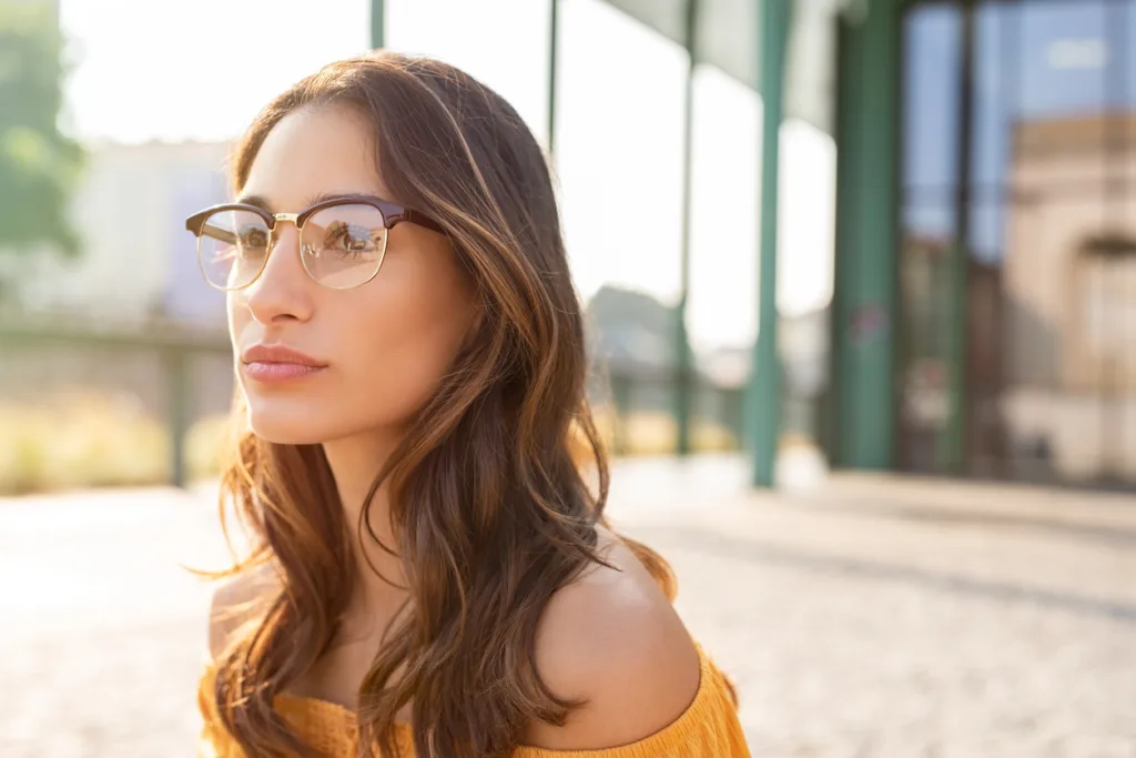 Portrait of trendy city girl with eyeglasses looking away. Young latin woman thinking and wearing spectacles. Outdoor closeup of thoughtful woman with urban background at sunset with copy space.