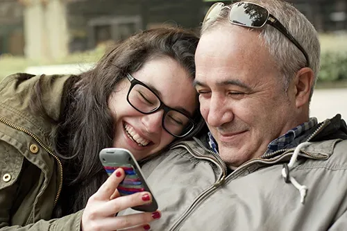 Father and teenager daughter with head in his shoulder sharing something funny in a mobile phone