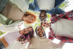 Low angle view of a group of multiracial happy young teenagers hugging each others in a circle, looking down at camera with smiling faces, representing friendship, diversity, and togetherness outdoors. High quality photo