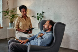 Frustrated middle age man sitting in armchair and solving his mental problems while having therapy session with african female psychologist