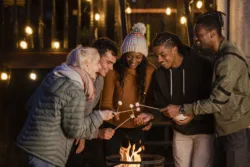 A group of young adults friends are standing around a fire in the dark, smiling. They are heating up the marshmallows they have on sticks.