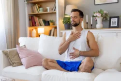 A man in white tank top and blue shorts sits on a white couch in his living room with hands on his chest, reflecting peacefully in an environment filled with calm and warmth.A man sits cross-legged on a white carpet with eyes closed, hands placed on his chest and stomach, wearing a white tank top and blue shorts in a decorated living room.