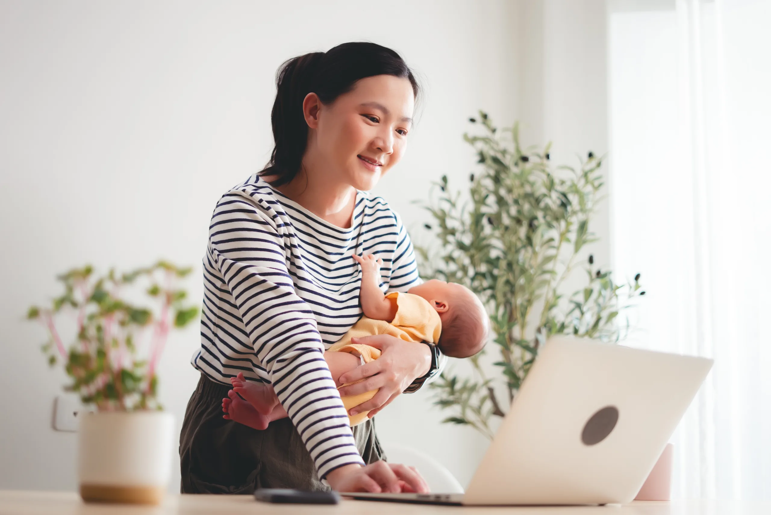 Asian woman carrying her baby while working with laptop at home office.