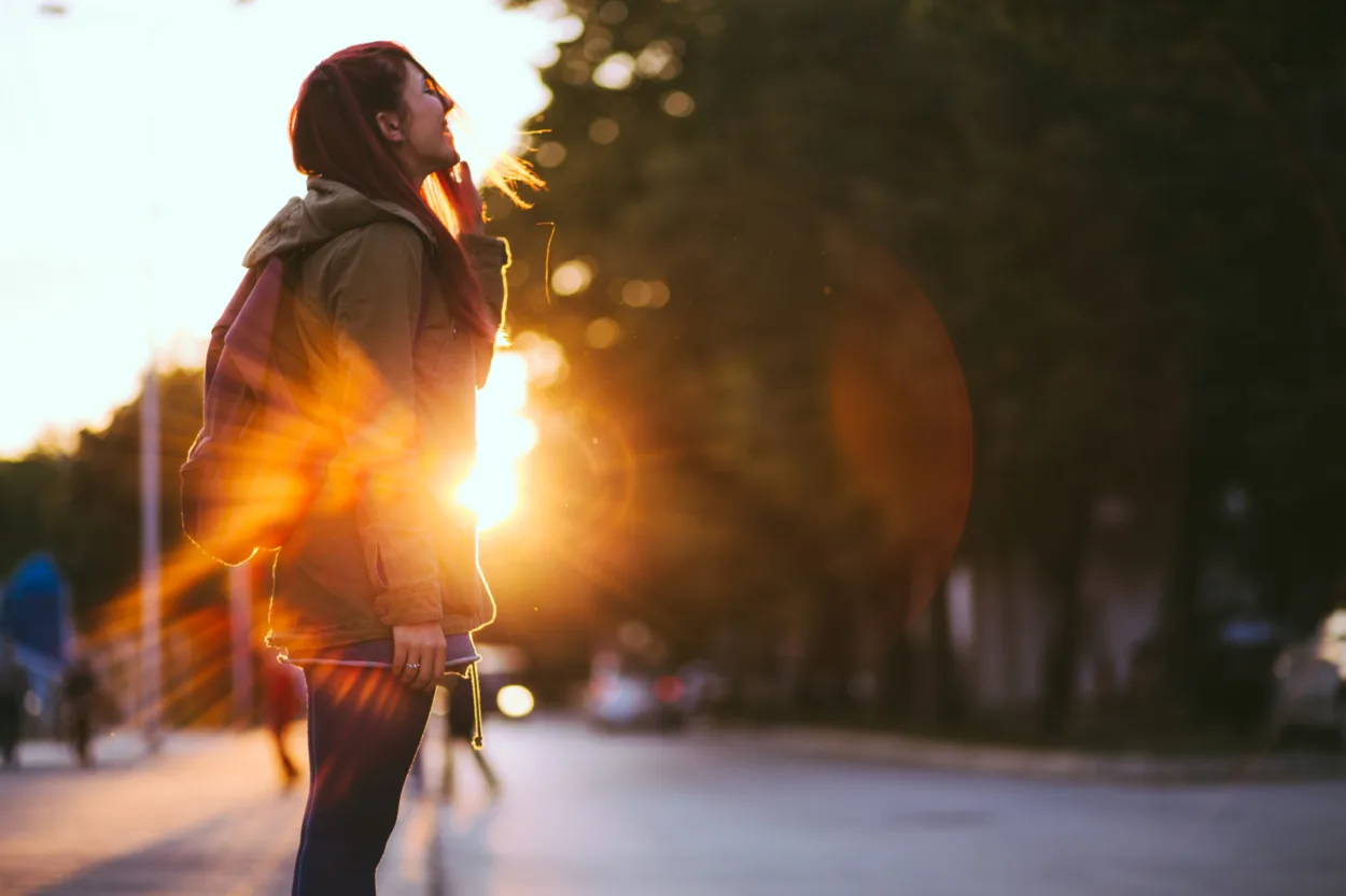 Smiling Young Red-haired woman dressed in jacket is enjoying vivid sunset in the streets.