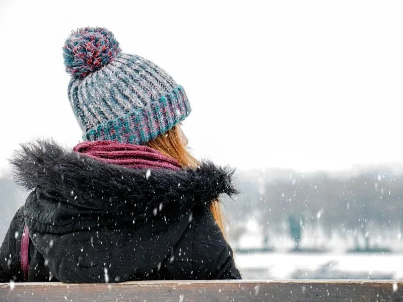 Depressed woman sits on bench on a snowy day, bundled up in hat, scarf, and coat