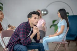 Worried young adult sitting with his back turned to the group in a session