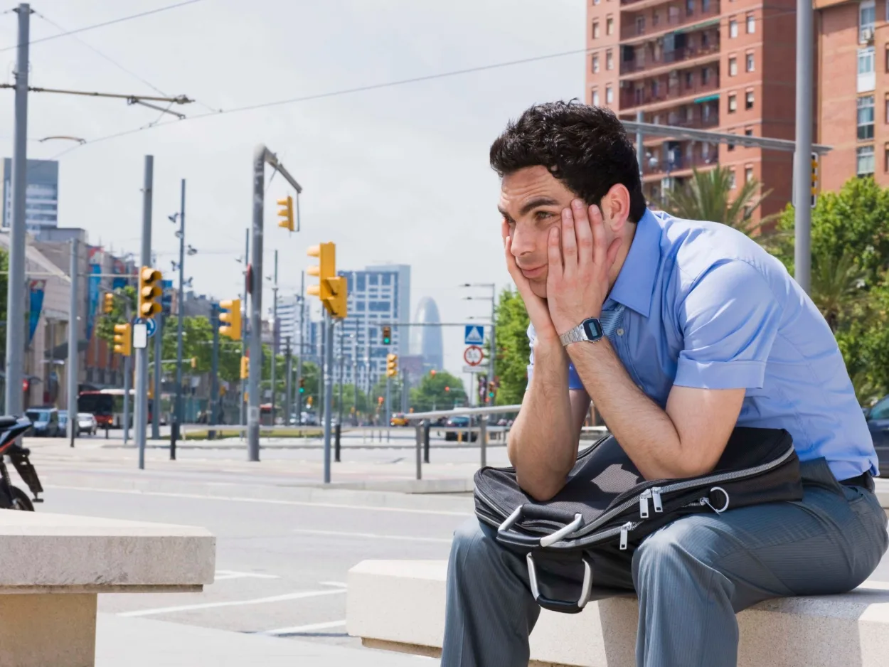 A sad man sits on a bench in a city with his head in his hands