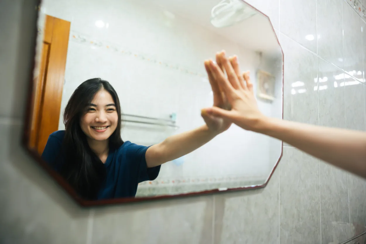 Young adult smile asian woman practice self talk conversation in the morning in bathroom at home. Hand hi-touch at mirror. Healthy lifestyle after wake up concept.