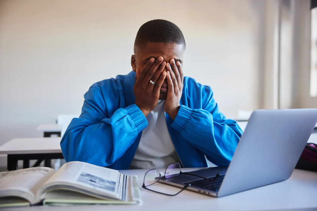 Young male college student looking stressed with his head in his hands while trying to do his homework at a table in a classroom at school