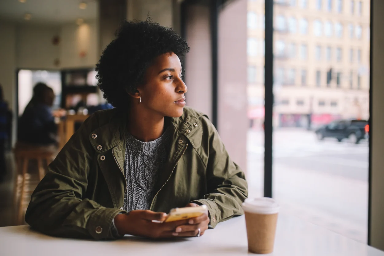 Young mixed race woman sitting at the desk in a cafe in downtown Los Angeles, texting at her smartphone while having a coffee.