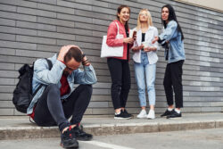 Three women standing and laughing at guy that sitting outdoors near building at daytime.