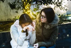 Two teenager girls, sisters, in the city square illuminated for Holidays. One girl crying and other sister comforting her.